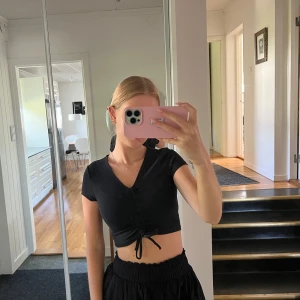 Svart croppad topp med knyt - Säljer en svart croppad topp med knyt framtill. Den har korta ärmar och en v-ringad design. Perfekt för sommarens alla fester eller bara en chill dag ute. Superbekväm och lätt att matcha med allt! Ny med prislapp kvar.