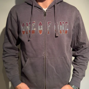 Grå hoodie med dragkedja från One of One - Säljer en snygg grå hoodie från One of One. Den har en dragkedja framtill och texten 'ONE OF ONE' i färgglada bokstäver på bröstet. Perfekt för en avslappnad stil och passar till alla tillfällen. Den är i bra skick och superbekväm! limited edition 1/100