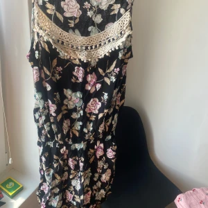 Blommig playsuit med spetsdetaljer - Säljer en superfin blommig playsuit med vackra spetsdetaljer vid halsen. Klänningen har en svart bas med rosa och vita blommor som ger en romantisk känsla. Perfekt för sommarens fester eller en dag på stan!