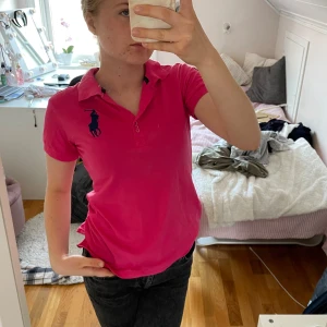 Ralph lauren  - Säljer en snygg rosa pikétröja med en stor mörkblå logga på bröstet. Tröjan har en klassisk krage och korta ärmar, perfekt för en casual look. Den är i bra skick och passar både till vardags och lite mer uppklädda tillfällen.