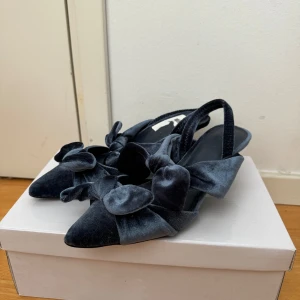 Svarta sammetsskor med rosett - Säljer ett par eleganta blå sammetsskor med spetsig tå och vacker rosett på ovansidan. Skorna har en liten klack och en smidig rem runt hälen för extra komfort. Perfekta för festliga tillfällen eller en stilfull vardag.
