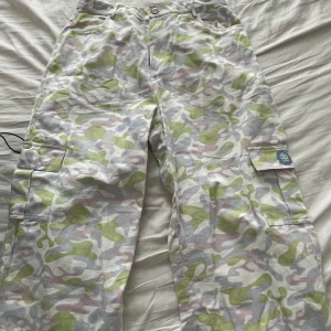 Cargobyxor med kamouflagemönster - Säljer ett par Bratz Limited edition cargobyxor med ett coolt kamouflagemönster i grönt, rosa och vitt. Byxorna har praktiska fickor på sidorna och en snygg broderad detalj med en blå logga. strl 40/L