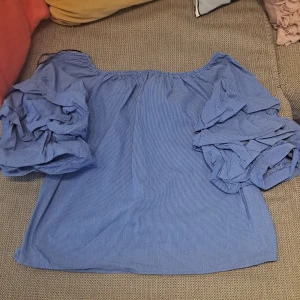 Blå randig offshoulder topp - Säljer en superfin blå offshoulder topp med randigt mönster. Den har stora, puffiga ärmar som ger en unik look. Perfekt för sommardagar eller en festlig kväll!