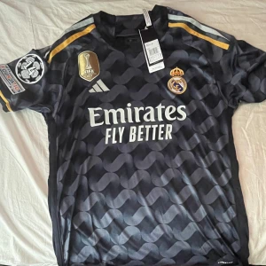 Real Madrid fotbollströja - Säljer en snygg Real Madrid fotbollströja från Adidas. Tröjan är svart med ett coolt mönster och har korta ärmar. Den har Real Madrids emblem broderat på bröstet och texten 'REAL MADRID' på baksidan. Det finns även FIFA-märket och Champions League-loggan på ärmen. Perfekt för alla Real Madrid-fans!