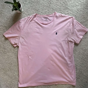 Rosa t-shirt från Polo Ralph Lauren - Säljer en stilren rosa t-shirt från Polo Ralph Lauren med det klassiska broderade logotypen på bröstet. T-shirten är kortärmad och tillverkad i mjuk bomull för en bekväm passform. Perfekt för en avslappnad look.