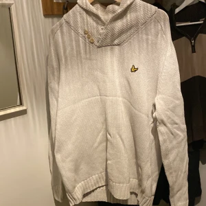 Vit stickad tröja med knappar - Säljer en vit stickad tröja från Lyle & Scott med en snygg krage och tre dekorativa knappar. Tröjan har ett broderat märke på bröstet och långa ärmar. Perfekt för kyligare dagar!