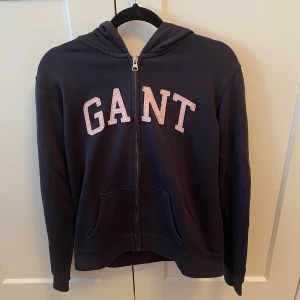 Mörkblå hoodie från GANT - Säljer en snygg mörkblå hoodie från GANT med dragkedja och stor logga i vitt på framsidan. Tröjan har en bekväm huva med ljus insida och praktiska fickor framtill. Perfekt för en avslappnad stil. Köpare står för frakten 🩷