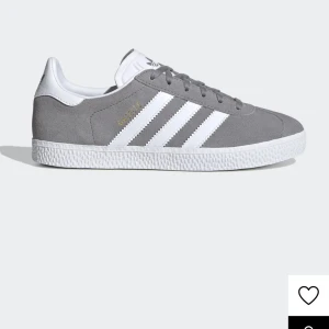 Grå Adidas Gazelle sneakers - Snygga grå Adidas Gazelle sneakers med klassiska vita ränder och snörning. Skorna har en stilren design med en vit sula och gyllene detaljer på sidan. Perfekta för en avslappnad stil.