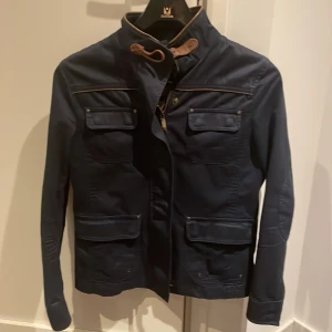 Blå jacka från Massimo Dutti - Snygg blå jacka från Massimo Dutti med stilrena detaljer. Jackan har långa ärmar och flera fickor med knappar. Den har en dragkedja framtill och en hög krage med bruna detaljer. Perfekt för en stilren look.