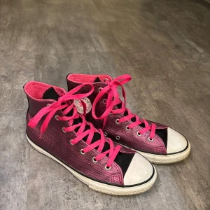 Rosa höga sneakers från Converse - Snygga rosa höga sneakers från Converse med häftig design. Skorna har en texturerad yta och vita gummisulor. De är utrustade med rosa snörning och den ikoniska Converse-loggan på sidan.