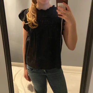Svart blus med volangdetaljer - Säljer en svart blus med volangdetaljer vid ärmarna. Blusen har en smockad detalj vid bysten och är ärmlös. Perfekt för en stilren look.
