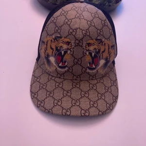 Gucci keps beige  - Snygg beige keps från Gucci med ikoniskt GG-mönster och coolt tigerbroderi framtill. Kepsen har en svart mesh-baksida för ventilation och ett justerbart band med röd-gröna detaljer. Perfekt för att ge din outfit en lyxig touch.