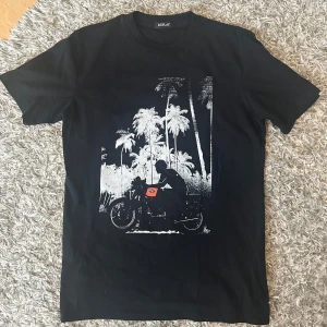 Replay T-Shirt - Snygg svart t-shirt från Replay med ett coolt tryck av en motorcykel och palmer i vitt och rött. Perfekt för en avslappnad stil med en touch av äventyr. T-shirten har en klassisk passform och är kortärmad. 