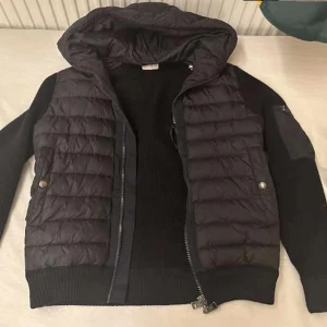 Moncler Cardigan - Snygg svart quiltad jacka från Moncler med huva och dragkedja. Jackan har långa ärmar och praktiska fickor med knappar. Perfekt för kyligare dagar. Priset redan prutat och klart. Går att fixa i alla storlekar