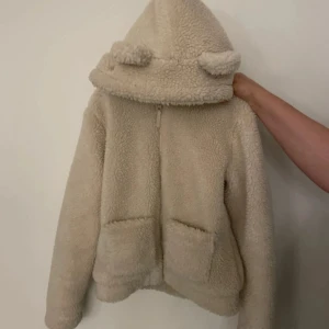 Beige teddyjacka med huva från H&M - Mysig beige teddyjacka från H&M med huva och dragkedja framtill. Jackan har långa ärmar och två fickor fram. Perfekt för kyliga dagar när du vill vara både varm och stilren.