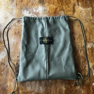 Stone Island Ryggpåse  - Snygg grön ryggpåse med dragsko och ett stilrent märke på framsidan. Väskan har en enkel design med två snören för att justera öppningen. Perfekt för en avslappnad stil. Finns ett fack med dragkedja inuti. Köpt som present till min son som inte tyckte den var tillräckligt cool 🥲