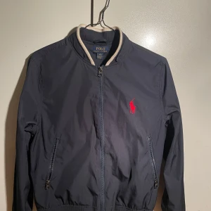 Blå jacka från Ralph Lauren - Säljer en stilren jacka från polo Ralph Lauren med dragkedja och ribbade detaljer vid krage, ärmslut och nederkant. Jackan har en broderad röd logotyp på bröstet och praktiska fickor med dragkedja. Perfekt för en casual look! Den är i bra skick men en liten fläck på ryggen som går bort i tvätten, det är storlek L 14-16. Men skulle säga att den passar xs😊kan sänka mig lite i pris vid snabb affär💯