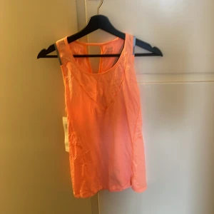 Orange linnetopp från Casall - Säljer en fräsch orange linnetopp från Casall. Perfekt för träning med sin ärmlösa design och bekväma passform. Toppen har en stilren look med grå detaljer vid axlarna.