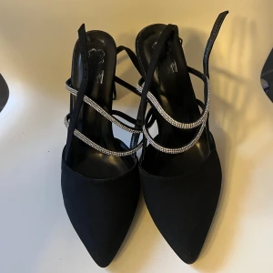 Svarta klackskor med strassdetaljer - Eleganta svarta klackskor med spetsig tå och glittrande strassdetaljer runt foten. Perfekta för en kväll ute eller en speciell tillställning. Tillverkade i Turkiet. Endast använd en gång. Smutsig på undersidan men märks inte eftersom det är på undersidan.