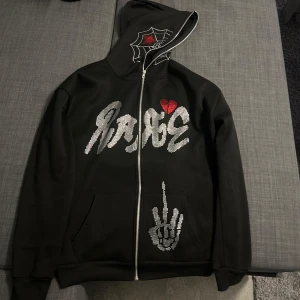 Svart hoodie med tryck - Snygg svart hoodie med dragkedja och coola detaljer gjorda av pärlor. Har ett stort tryck med text och ett rött hjärta på bröstet. På huvan finns ett spindelnätsmönster. Perfekt för en edgy look. Man kan dra upp dragkedjan hela vägen upp om man vill.