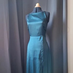 Mörkgrön långklänning med omlott - Har använts 3 gånger och har även syt upp lite nere vid kjolen med så få stygn som möjligt. Jag kan ta bort dem vid efterfrågan. Vill också påpeka att tyget är lätt skrynkligt.