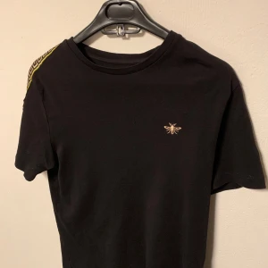 Svart t-shirt med gulddetalj - Snygg svart t-shirt med en liten gulddetalj i form av en insekt på bröstet. T-shirten har korta ärmar och en dekorativ rand i gult längs axeln. Perfekt för en stilren look. Storlek M.