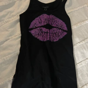 Svart linnetopp  Cyberdog - Cool svart linnetopp från Cyberdog med ett stort lila läpptryck på framsidan. Perfekt för en unik och edgy stil. Toppen är ärmlös och har en rund halsringning.