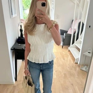 Vit ärmlös blus - Snygg vit ärmlös blus med volangdetaljer i midjan. Perfekt för en somrig look. Blusen har en smickrande passform och är lätt att matcha med jeans eller kjol.
