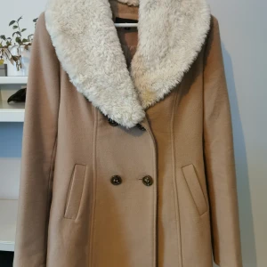 Beige kappa med fuskpäls från Tally Weijl - Snygg beige kappa från Tally Weijl med mjuk fuskpäls på kragen. Dubbelknäppt design med två fickor framtill. Perfekt för kyliga dagar när du vill hålla stilen.