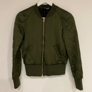 Olivgrön bomberjacka från H&M - Snygg olivgrön bomberjacka från H&M med quiltade ärmar och dragkedja framtill. Jackan har en klassisk bomberstil med ribbade muddar vid ärmslut och nederkant. Perfekt för en cool och avslappnad look. OBS Tyvärr har jackan en stor fläck på ryggen, vet ej vad det är då den hängt i garderoben i flera år. Det kanske går att tvätta bort men jag vet inte då jag inte har testat.