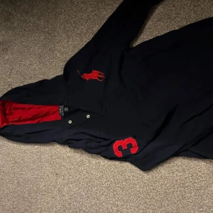 Svart pikétröja från Ralph Lauren - Snygg svart pikétröja från Ralph Lauren med röd insida på kragen och en stor röd logga på bröstet. Tröjan har knappar framtill och är tillverkad i 100% bomull. Perfekt för en stilren look. Strlk 170