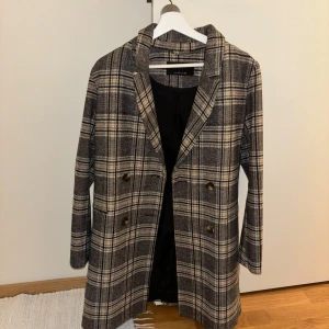 Rutig kappa från Sinsay - Snygg rutig kappa från Sinsay i storlek M. Den har en klassisk krage och knappar framtill. Perfekt för höst och vår med sitt stilrena mönster i jordnära färger.