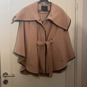 Beige kappa/poncho från Stockh LM - Elegant beige kappa från Stockh LM med svarta kantdetaljer och knappar. Den har en stilren design med bälte i midjan och en vid passform som ger en chic look. Perfekt för höst och vår.