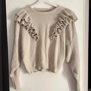 Beige tröja med volangdetaljer - Säljer en mysig beige tröja med volangdetaljer på framsidan. Tröjan har långa ärmar och en rund halsringning. Perfekt för en stilren och bekväm look.