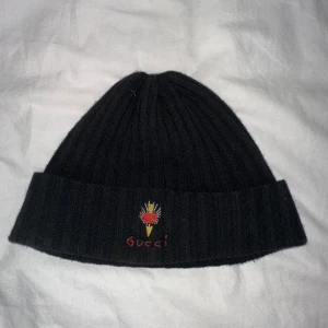 Svart ribbad mössa från Gucci - Snygg svart ribbad mössa från Gucci med broderad detalj framtill. Perfekt för att hålla värmen med stil under kyliga dagar. Tillverkad i Italien. Kvitto finns