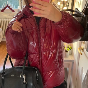 Vinröd pufferjacka - Snygg vinröd pufferjacka med glansig finish och dragkedja framtill. Jackan har långa ärmar och en hög krage för extra värme. Perfekt för kyliga dagar. Väldigt lik den virala Nelly jackan! 🌟Pris går att diskutera 