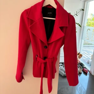 Rosa kappa från Promiss - Snygg rosa kappa från Promiss med knappar och knytskärp i midjan. Jackan har långa ärmar och en klassisk krage. Perfekt för att ge en färgklick till din outfit!