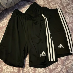 Svarta träningsshorts från Adidas - Säljer ett par svarta träningsshorts från Adidas med de klassiska tre vita ränderna längs sidorna. Shortsen har elastisk midja och är tillverkade i ett lätt och bekvämt syntetmaterial, perfekt för sport och träning. För dom var små på mig