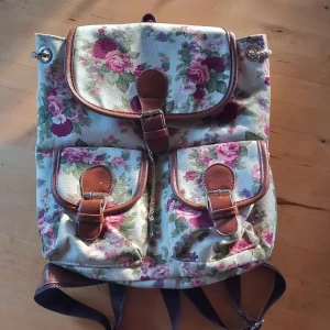 Blommig ryggsäck med läderdetaljer - Charmig ryggsäck med blommigt mönster och snygga läderdetaljer. Ryggsäcken har flera fickor och justerbara remmar för bekväm användning. Inga hål eller defekter. 