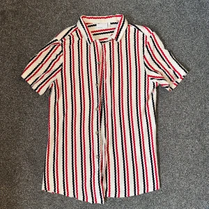 Randig skjorta från ASOS - Snygg skjorta från ASOS med korta ärmar och ett randigt mönster i rött, svart och vitt. Skjortan har knappar framtill och är perfekt för en stilren look.