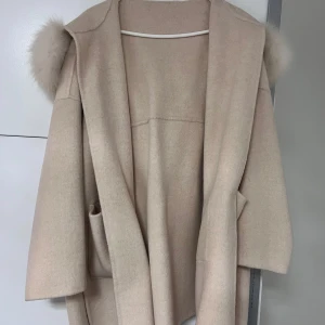 Beige kappa med pälsdetaljer - Elegant beige kappa med stor huva och pälsdetaljer. Kappan har ett knytbälte i midjan och praktiska fickor framtill. Perfekt för kyliga dagar när du vill hålla stilen. Äkta päls. Obs:Kappan är ljusare än vad den ser ut (bild 3 visar mer riktiga färgen) 