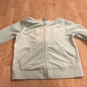 Ljusblå hoodie från Juicy Couture - Mysig ljusblå hoodie från Juicy Couture med dragkedja och känguruficka. Den har en broderad logga på bröstet och justerbar huva med dragsko. Perfekt för en avslappnad stil.