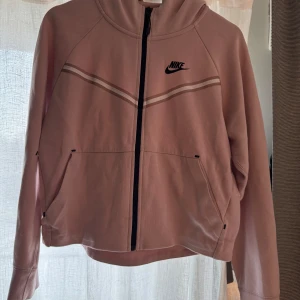 Rosa hoodie från Nike - Säljer en rosa hoodie från Nike med dragkedja och huva. Den har ett stilrent mönster med svarta detaljer och fickor framtill. 