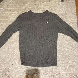 Grå kabelstickad tröja från Polo Ralph Lauren - Säljer en stilren grå stickad tröja från Polo Ralph Lauren med kabelstickat mönster. Tröjan har långa ärmar och en klassisk rund halsringning. Perfekt för kyligare dagar. Storlek L Barn