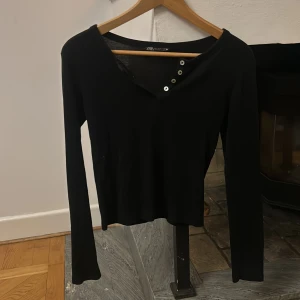 Svart långärmad topp från Zara - Snygg svart långärmad topp från Zara med knappar vid halsringningen. Sitter väldigt fint på. Säljer då den är lite för liten. Storlek s. Har ett väldigt mjukt material och sitter ganska tajt.