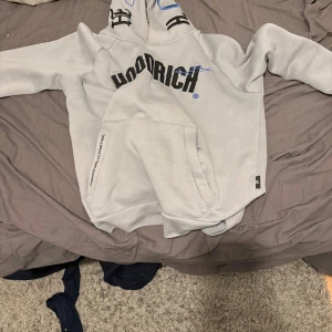 Grå hoodie från Hoodrich - Säljer en grå hoodie från Hoodrich med svart text på framsidan och tryck på baksidan. Den har en stor ficka framtill och en justerbar huva. Perfekt för en avslappnad stil.