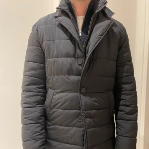 Svart jacka från John Henric - Snygg svart quiltad jacka från John Henric med avtagbar huva. Jackan har både dragkedja och knappar framtill. Inbyggd väst som är avtagbar.