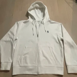 Vit hoodie från Polo Ralph Lauren - Säljer en stilren vit hoodie från Polo Ralph Lauren med dragkedja och klassisk logga på bröstet. Perfekt för en avslappnad look. Den har en justerbar huva och praktiska fickor framtill. Använd ett par gånger men inga täcken på användning förutom att den är lite nopprig men inget man tänker på.