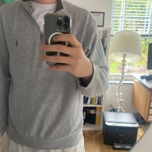 Grå tröja med dragkedja från Ralph Lauren - Säljer en stilren grå tröja från Ralph Lauren med halv dragkedja och broderad logga på bröstet. Perfekt för en avslappnad och snygg look. Tröjan har långa ärmar och är tillverkad i mjukt material. Nästan aldrig använd så skick är 9/10 