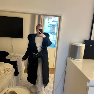 Svart lång dunjacka med pälskrage - Snygg och varm svart lång dunjacka med en fluffig pälskrage. Perfekt för kalla vinterdagar. Jackan har knappar så man kan stänga den och ett snöre man kan knyta som man vill runt midjan. Använd en gång, i helt nytt skick och köpt för 2000:-, Inge defekter!